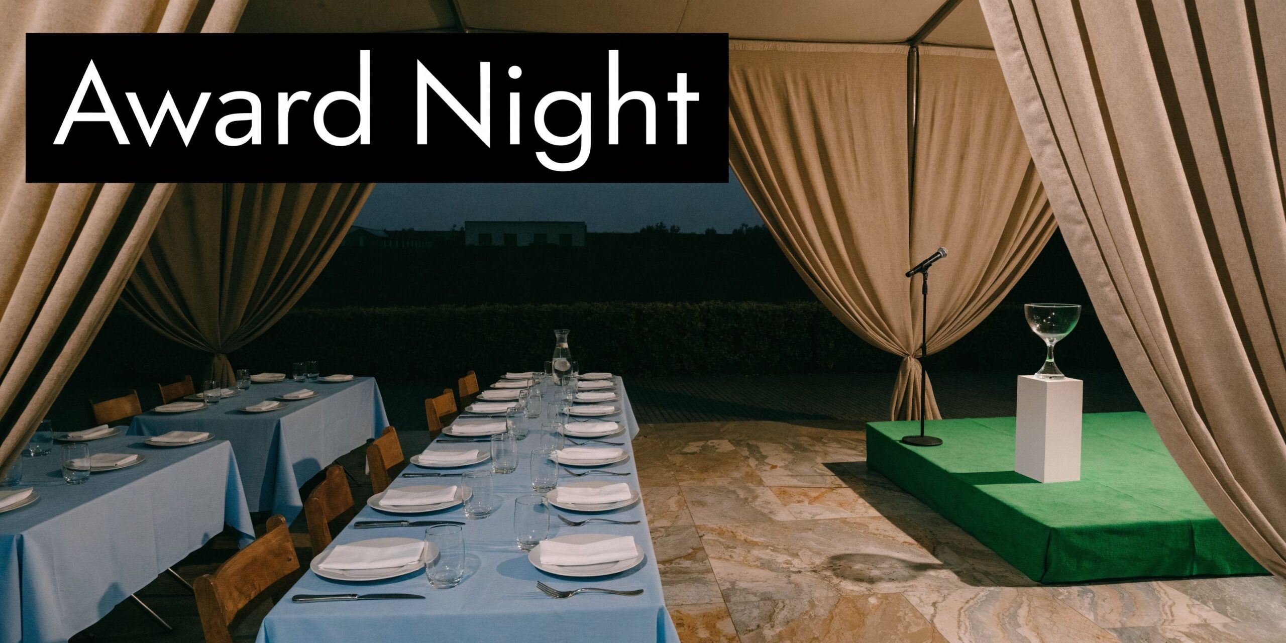 A formal event setup outdoors with blue-covered tables and a stage featuring a trophy and microphone.