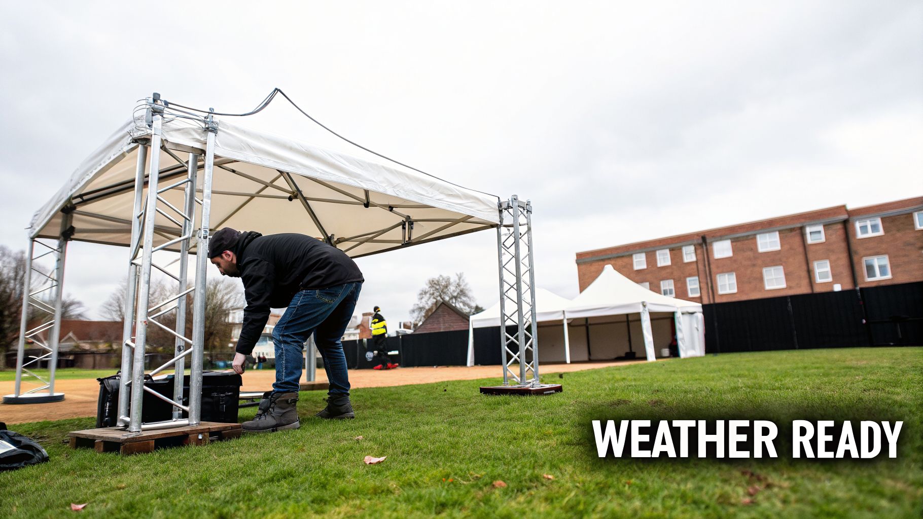 A person sets up a white gazebo structure on a grassy field with other tents and a building.