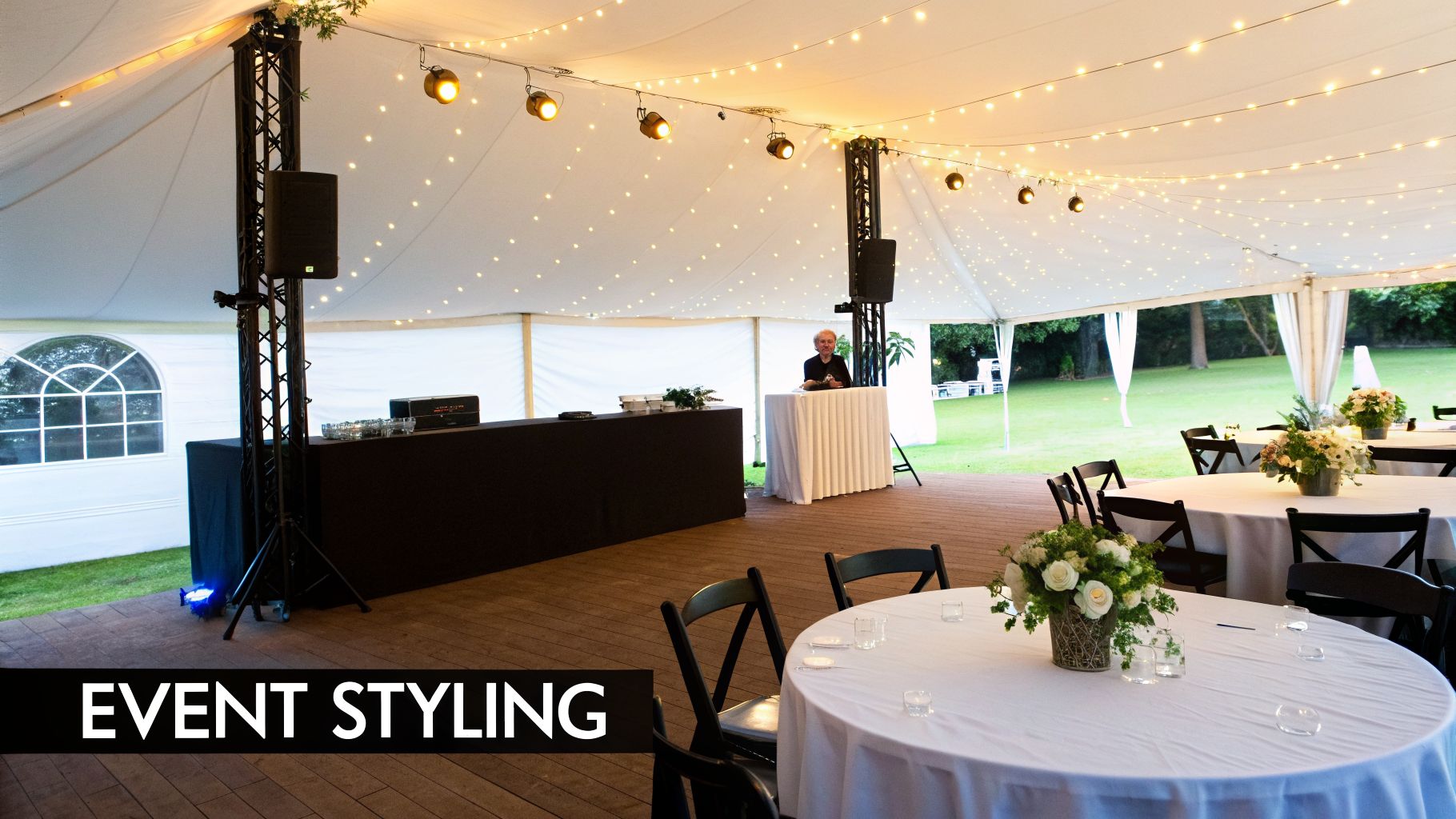 A spacious white marquee tent prepared for an elegant event, featuring table settings, a DJ, and string lights.