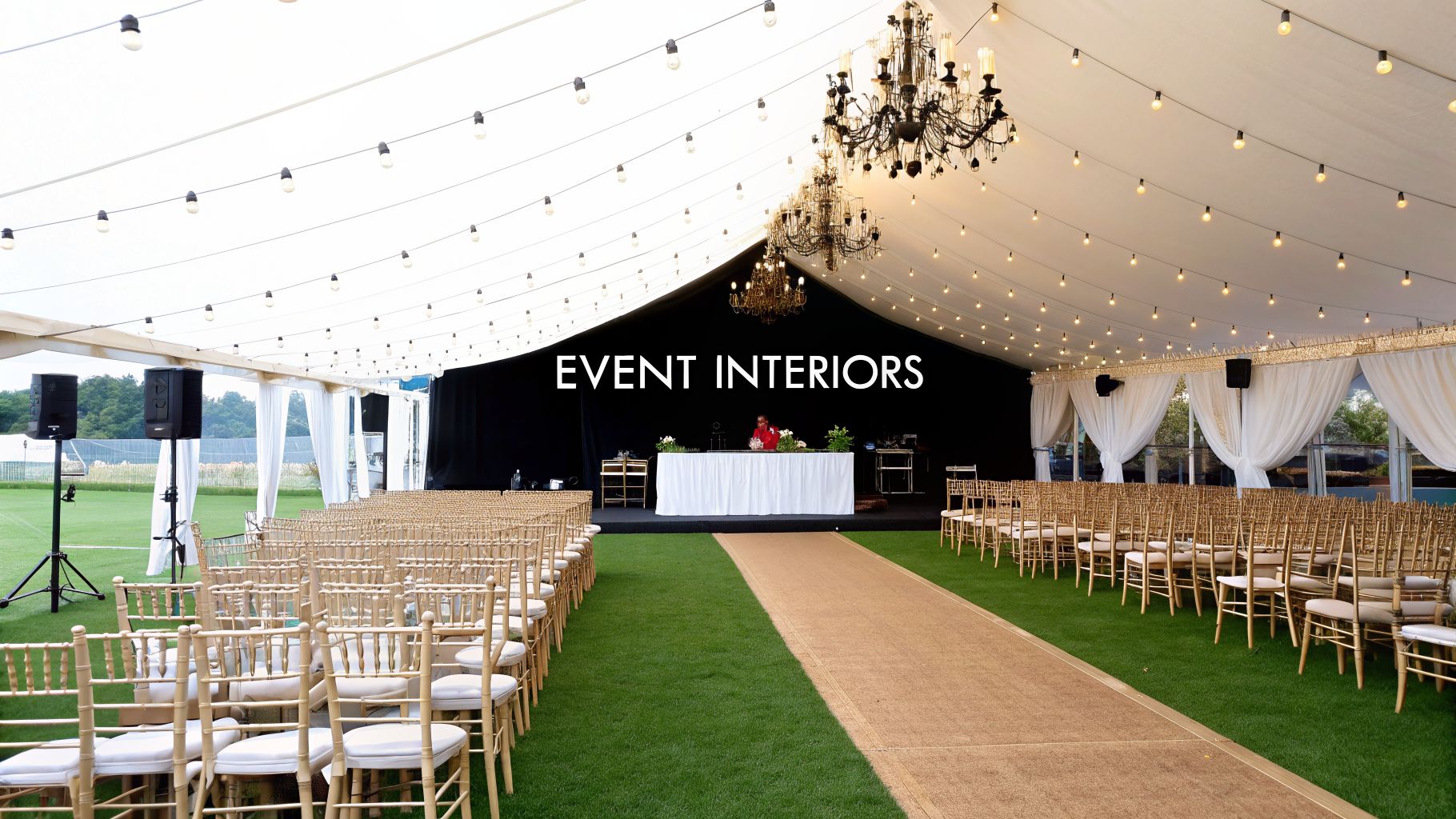 Inside an event marquee featuring gold chairs, a brown aisle, stage, and elegant lighting.