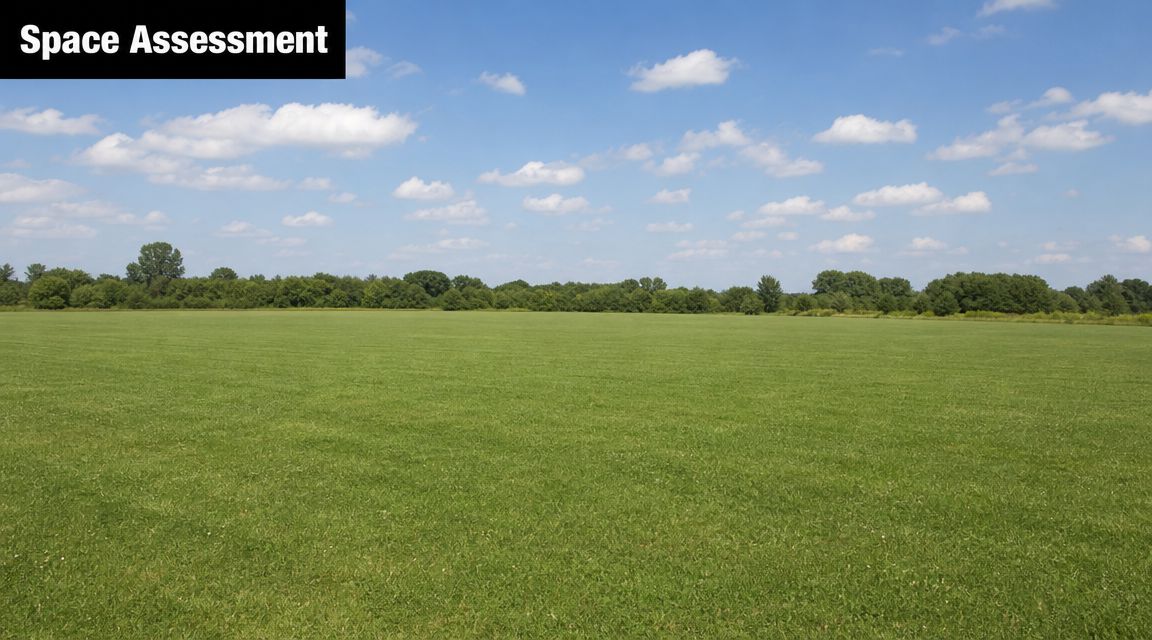 A wide, open green field under a clear blue sky with a line of trees in the distance.